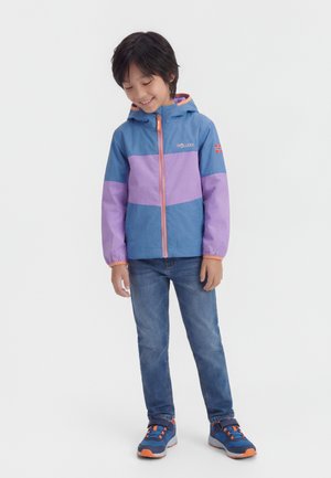 Bambino sorridente con indosso una giacca con cappuccio blu e viola, jeans blu e scarpe da ginnastica blu con dettagli arancioni, in piedi su uno sfondo bianco.