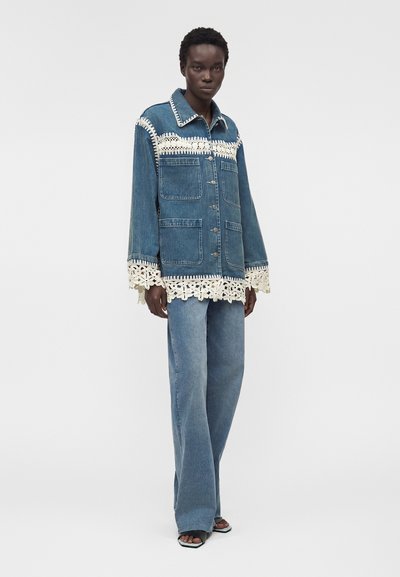 Μοντέλο φοράει μπλε τζιν μπουφάν με εκρού κροσέ λεπτομέρειες στον γιακά, τα μανίκια και το τελείωμα, συνδυασμένο με φαρδιά μπλε τζιν παντελόνια και μαύρα ανοιχτά παπούτσια.