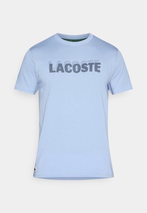 Světle modré tričko s krátkými rukávy z bavlny. Na hrudi má velké černé logo "LACOSTE". Standardní střih s kulatým výstřihem.