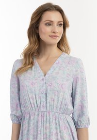Rochie florală de culoare albastru deschis, cu flori violete și albe. Dispune de un decolteu în V, închidere cu nasturi și talie strânsă cu mâneci bufante.