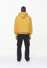 Gelber Kapuzenpullover mit großer Kapuze und lockerer Passform, kombiniert mit schwarzen Cargohosen mit Taschen und weißen Sneakers. Schlichter Hintergrund.