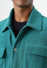 Groene wollen overhemdjas met een gestructureerde afwerking, voorzien van een puntige kraag, twee borstzakken en een zilveren knoopsluiting.