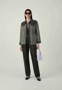 Kvinne kledd i svart og hvitt hundetannmønsterjakke med denimskuldre, svarte bukser, svarte sko, solbriller, og bærer en liten lavendelhåndveske.