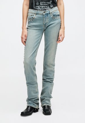 Jasnoniebieskie jeansy z denimu z srebrnymi ćwiekami w kształcie piramid na przednich kieszeniach i szwach bocznych, noszone z czarnymi butami na kwadratowym nosku.