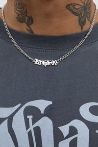 Colar de corrente prateada com um pêndulo metálico apresentando uma escrita curva intricada, usado sobre uma t-shirt cinza escura.
