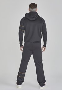 Zwarte sportieve trainingsoutfit met een capuchon. Bevat gouden accenten op de mouwen en de broeken. Gemaakt van gladde stof. Model is van achteren bekeken.