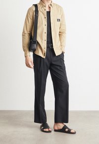 Béžová košile na zapínání s náprsní kapsou, černé kalhoty a černé nazouvací sandály. Doplňky zahrnují černou kabelku přes rameno. Hladká textura látky.