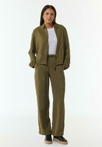 Olivegrüne Jacke und passende Hose aus glattem Stoff, mit elastischen Bündchen und verstellbarem Taillenbund; kombiniert mit schwarzen Sneakers.