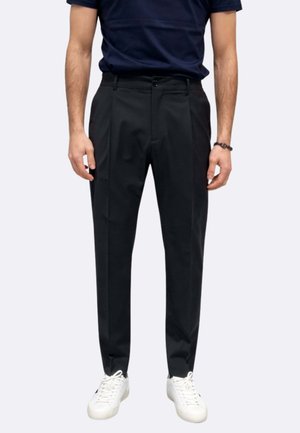 Uomo che indossa pantaloni neri su misura, t-shirt blu navy, sneakers bianche e un braccialetto nero al polso, in piedi davanti a uno sfondo semplice.