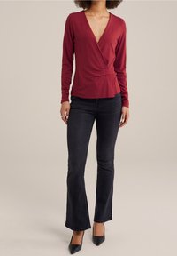 Woman wearing a fitted long-sleeve burgundy wrap top, dark bootcut jeans, black pointed-toe heels, and a silver ring on her left hand.