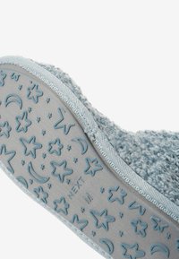 Zapatilla azul de felpa con una suela gris texturizada, que presenta un patrón de estrellas y lunas crecientes. Material suave y detalles en el borde visibles.