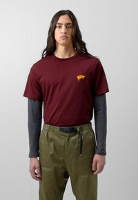 Camiseta corta de manga maroon con logo amarillo en el pecho, superpuesta a mangas largas grises. Pantalones verde oliva con bolsillos laterales y cinturón negro.