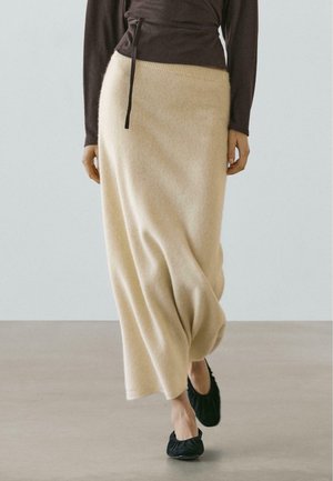 Gonna lunga color beige in misto lana con fascia elasticizzata in vita e pieghe delicate, abbinata a una maglia scura a maniche lunghe e scarpe basse nere.