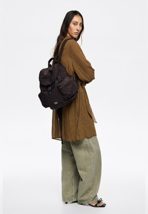 Mochila de nailon negra con parte superior redondeada, gran bolsillo delantero, tirantes ajustables y detalle sutil de logotipo. Lleva puesta sobre una blusa texturizada de color marrón claro y pantalones verdes.