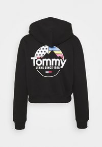 Sudadera corta negra con capucha. Presenta un logo circular multicolor en la parte posterior, que muestra montañas y estrellas con el texto "Tommy Jeans Desde 1985."