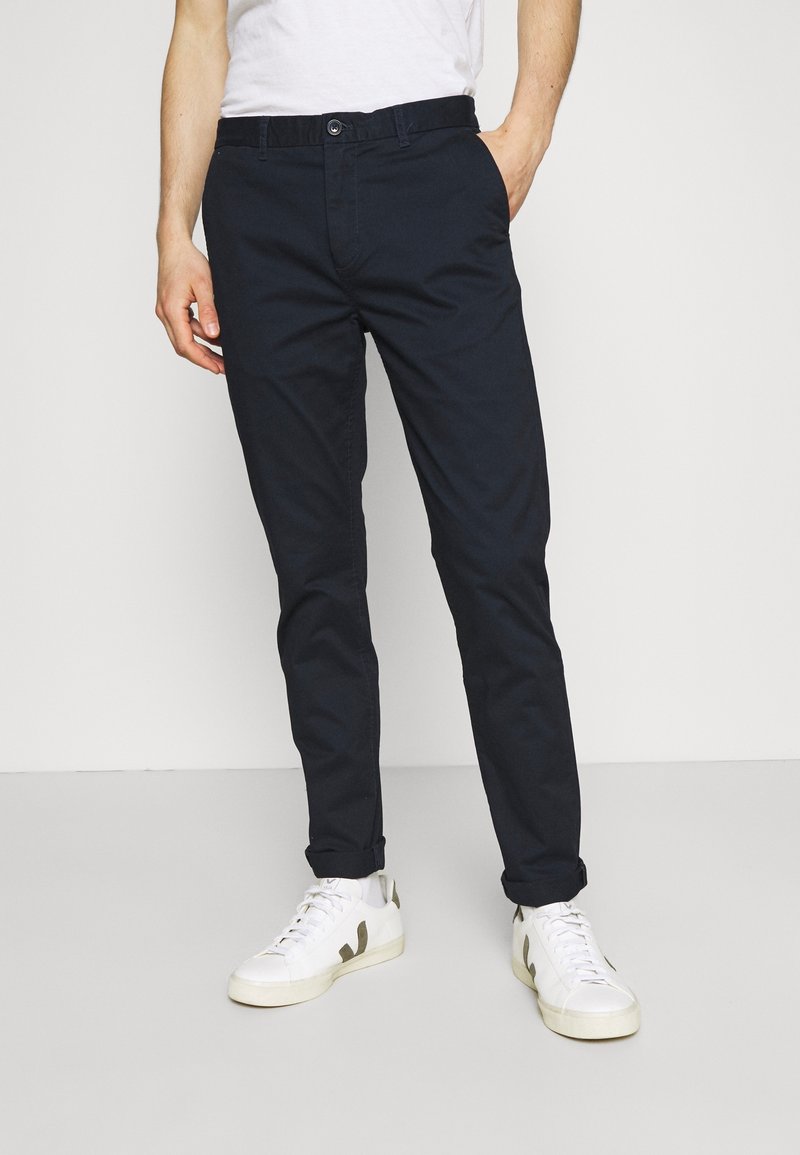 Chinos blu scuro in cotone, con vestibilità dritta, passanti per cintura e tasche frontali, abbinati a sneakers bianche con dettagli verdi.