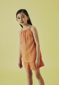 Completo due pezzi arancione con texture, composto da una canotta senza maniche e pantaloni corti, su uno sfondo giallo. Design semplice con una vestibilità rilassata.