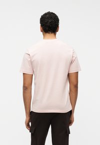 Homme aux cheveux bouclés portant un t-shirt à manches courtes rose clair et un pantalon cargo sombre, debout de dos sur un fond uni.
