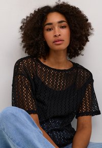 Schwarzes, offen gestricktes Oberteil mit kurzen Ärmeln, rundem Ausschnitt und gewelltem Saumdetail. Kombiniert mit hellen Jeans.