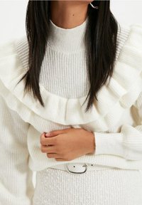 Weißer Pullover mit gerippter Textur und geschichteter Rüsche am Ausschnitt. Die Hand des Models liegt auf einem weißen Gürtel über einem strukturierten Rock.