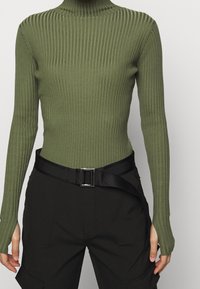 Olijfgroen geribbeld coltrui met lange mouwen, gecombineerd met zwarte high-waisted broek met een brede stoffen riem en zijzakken.