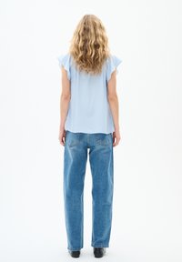 Blusa azzurra chiara a maniche corte con bordi arricciati, abbinata a jeans in denim chiaro e dal taglio morbido. L'outfit presenta un design rilassato.