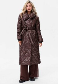 Brązowy pikowany długi płaszcz z wysokim kołnierzem, z paskiem w talii i ukośnym wzorem w romby, zestawiony z szerokimi brązowymi spodniami oraz czarnymi botkami.
