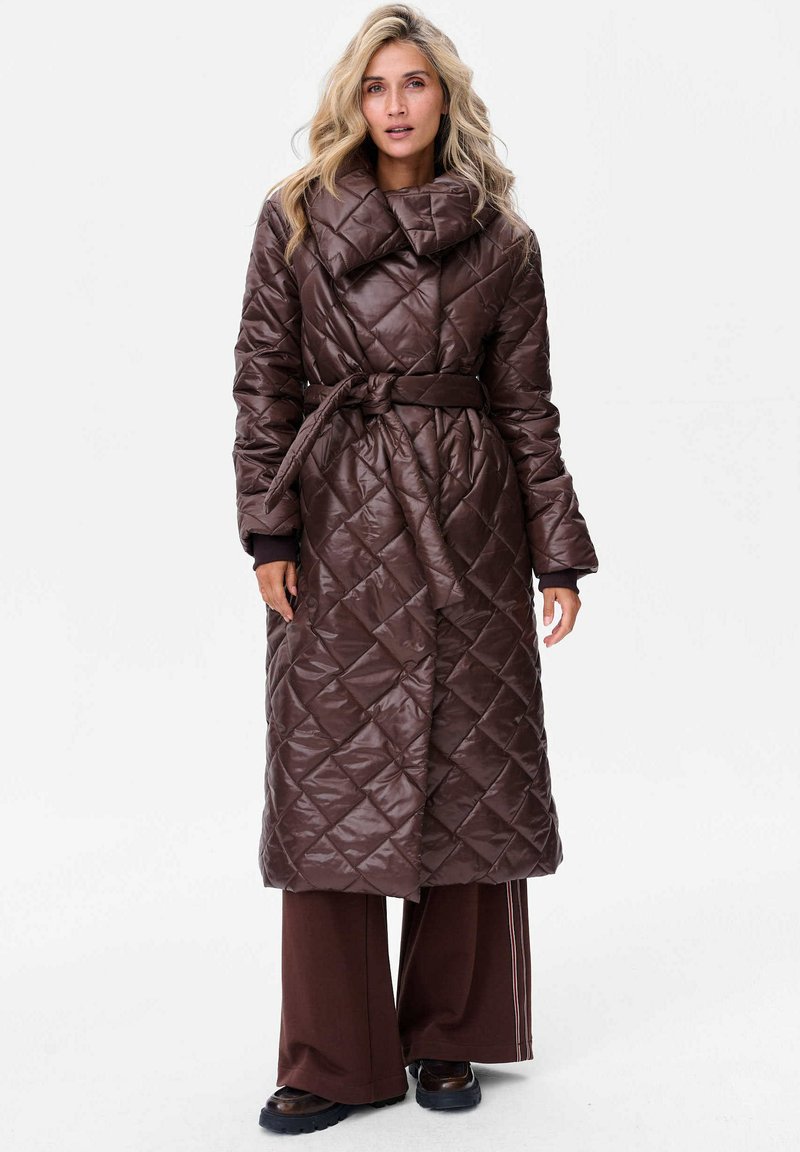 Brązowy pikowany długi płaszcz z wysokim kołnierzem, z paskiem w talii i ukośnym wzorem w romby, zestawiony z szerokimi brązowymi spodniami oraz czarnymi botkami.