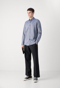 Light blue button-up shirt with long sleeves, black trousers, and black sneakers, held with a black clutch in a neutral background.