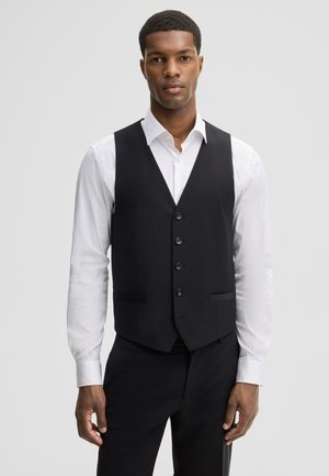 Homme portant un gilet noir boutonné sur une chemise blanche et un pantalon noir, debout devant un fond gris clair uni.