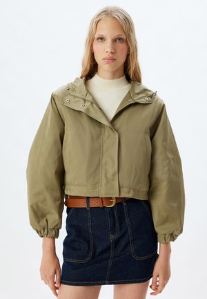 Affaldet olivengrønt jakke med hætte, har lynlås og knaplukninger, puffede ærmer og elastiske manchetter; båret over en lys top og denimnederdel.