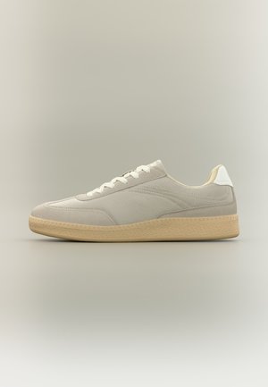 Baskets basses beiges en tissu texturé, détails en daim, lacets blancs, languette de talon blanche et semelle en caoutchouc marron clair sur fond neutre.