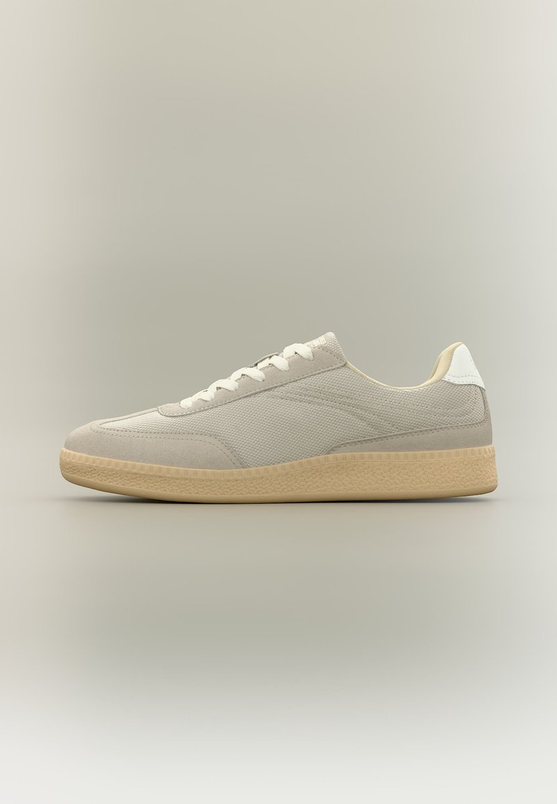 Μπεζ χαμηλού προφίλ αθλητικό παπούτσι με υφή υφάσματος, σουέτ λεπτομέρειες, λευκά κορδόνια, λευκή ταμπέλα στη φτέρνα και ελαφρώς καφέ λαστιχένια σόλα σε ουδέτερο φόντο.