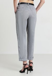 Persona che indossa pantaloni grigi con risvolti, con tasche laterali e posteriori, una maglietta nera e sandali con tacco alto neri, in piedi su un pavimento chiaro.