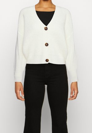 Personne portant un cardigan en grosse maille blanc avec trois grands boutons sur un haut noir et un pantalon noir sur un fond uni.