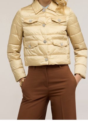Beige quiltet jakke med dekorative krystal knapper båret med højtaljede brune bukser, hænderne i lommerne og guldbøjler.