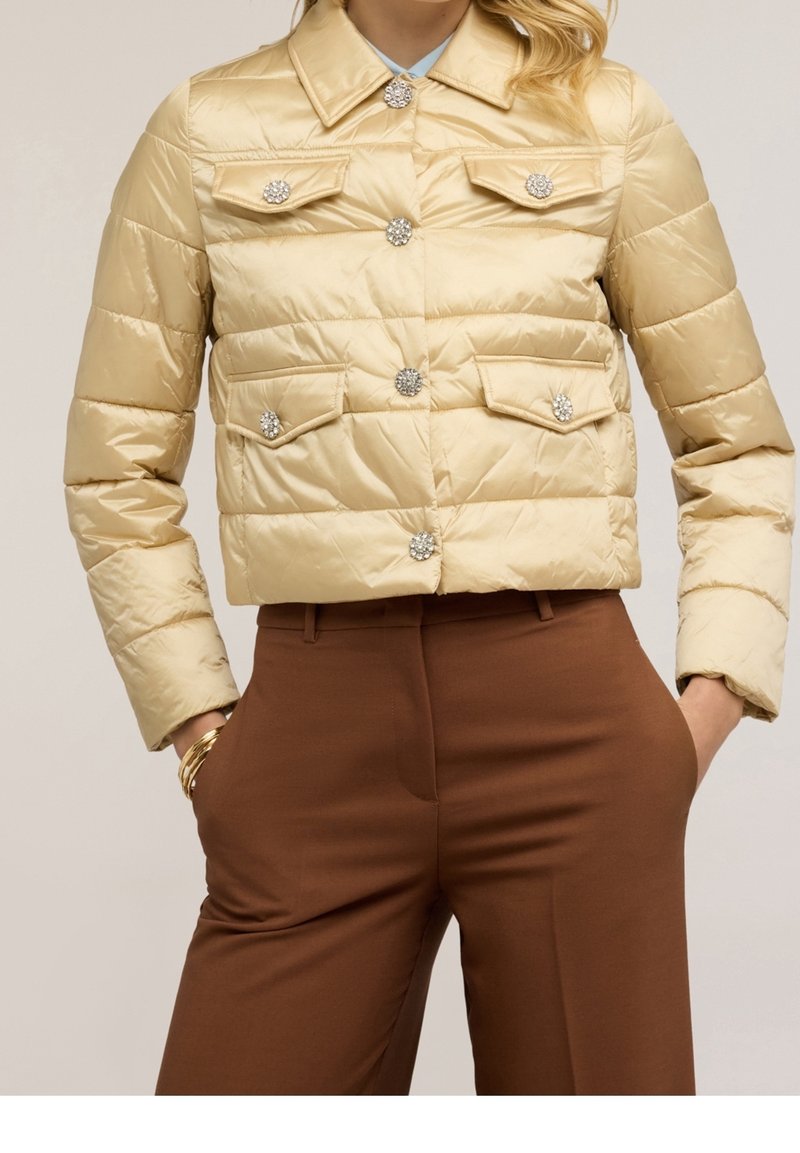 Veste matelassée beige avec des boutons en cristal décoratifs portée avec un pantalon marron taille haute, mains dans les poches et bracelets dorés.
