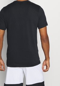 Hombre vestido con una camiseta negra básica de manga corta y shorts blancos con paneles negros, visto desde atrás sobre un fondo claro y sencillo.