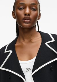 Czarna marynarka z białym wykończeniem, z klapami i dwoma guzikami. Pod spodem biała, prążkowana bluzka na ramiączkach z metalicznym logo.