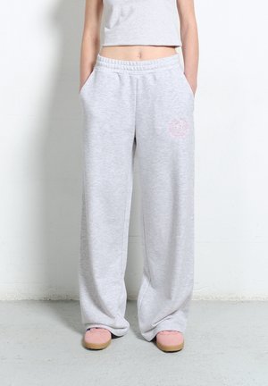 Osoba w luźnych, jasnoszarych dresach z rękami w kieszeniach, białym krótkim topie i różowych butach, stojąca na tle jednolitej ściany.