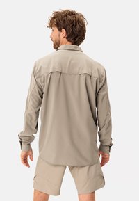 Camicia khaki a maniche lunghe in tessuto leggero con vestibilità rilassata, caratterizzata da un pezzo posteriore e maniche arrotolabili. Abbinata a pantaloni corti beige.