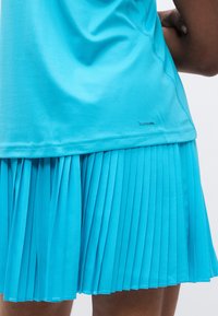 Ένα αθλητικό τοπ σε ανοιχτό γαλάζιο χρώμα με λείο ύφασμα που μετατρέπεται σε πλισέ φούστα στο ίδιο μπλε, με λογότυπο κοντά στο τελείωμα.