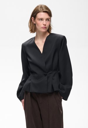 Vrouw met kort lichtbruin haar, draagt een zwarte blazer met één knoop en donkerbruine geplooide broek tegen een effen witte achtergrond.