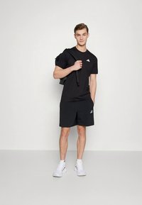 T-shirt e calções desportivos pretos, ambos com detalhes em logótipo. Modelo segura uma bolsa preta e calça ténis brancos. Design simples e ajustado.