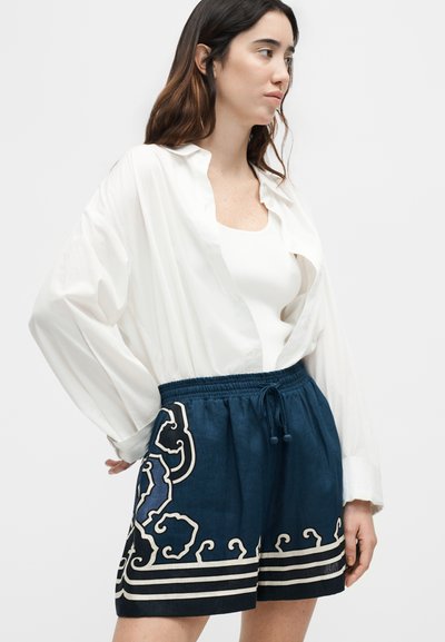 Γυναίκα με μακριά καστανά μαλλιά που φοράει ένα φαρδύ λευκό πουκάμισο, λευκή μπλούζα και σκούρα μπλε σορτς με λευκά αφηρημένα σχέδια, ποζάροντας μπροστά σε ένα απλό φόντο.