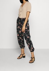 Beiger Blusen-Top mit kurzen Ärmeln, kombiniert mit schwarzen, floral gemusterten Hosen mit pinken Blumen und elastischen Bündchen. Schwarze Sandalen mit Knöchelriemen.