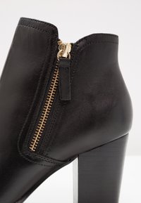 Bottine en cuir noir avec fermeture éclair dorée et talon bloc sur fond blanc.