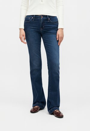 Άτομο που φοράει σκουρό μπλε τζιν bootcut, λευκό πουκάμισο με μακριά μανίκια και καφέ επίπεδα δερμάτινα παπούτσια με κορδόνια, στέκεται μπροστά σε ένα απλό φόντο.