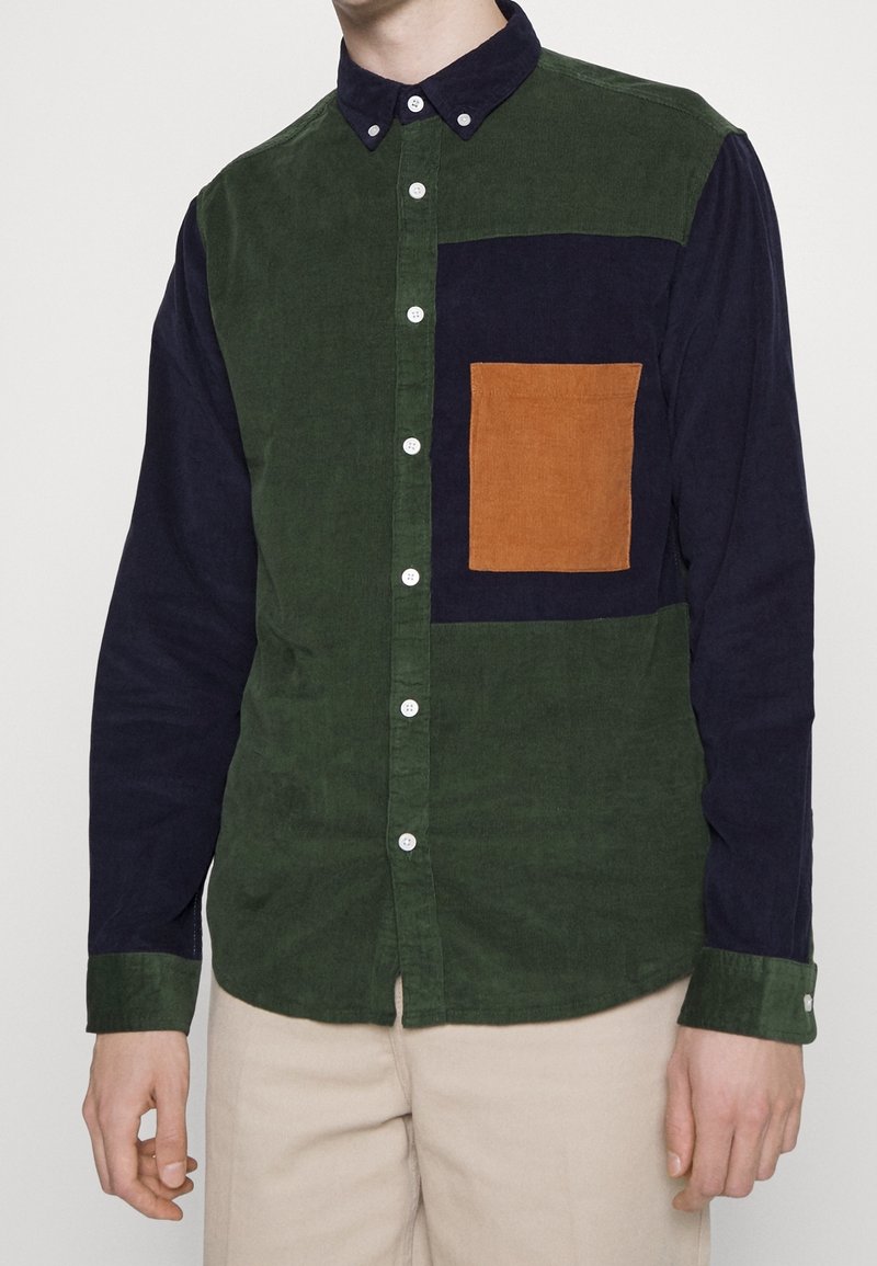 Knapp-up skjorta med grön och marinblå kordtyg, med en brun rektangulär ficka, vita knappar och kontrasterande ärmar.