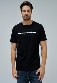 Hombre con cabello oscuro vistiendo una camiseta negra con el texto "SALSA JEANS" en el pecho y jeans azules, de pie contra un fondo gris liso.